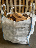 A large white woven bulk bag with blue trim and white handles, filled with cut firewood logs, photographed indoors against a wooden pallet background.