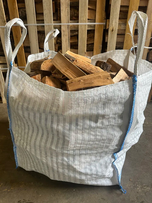 A large white woven bulk bag with blue trim and white handles, filled with cut firewood logs, photographed indoors against a wooden pallet background.